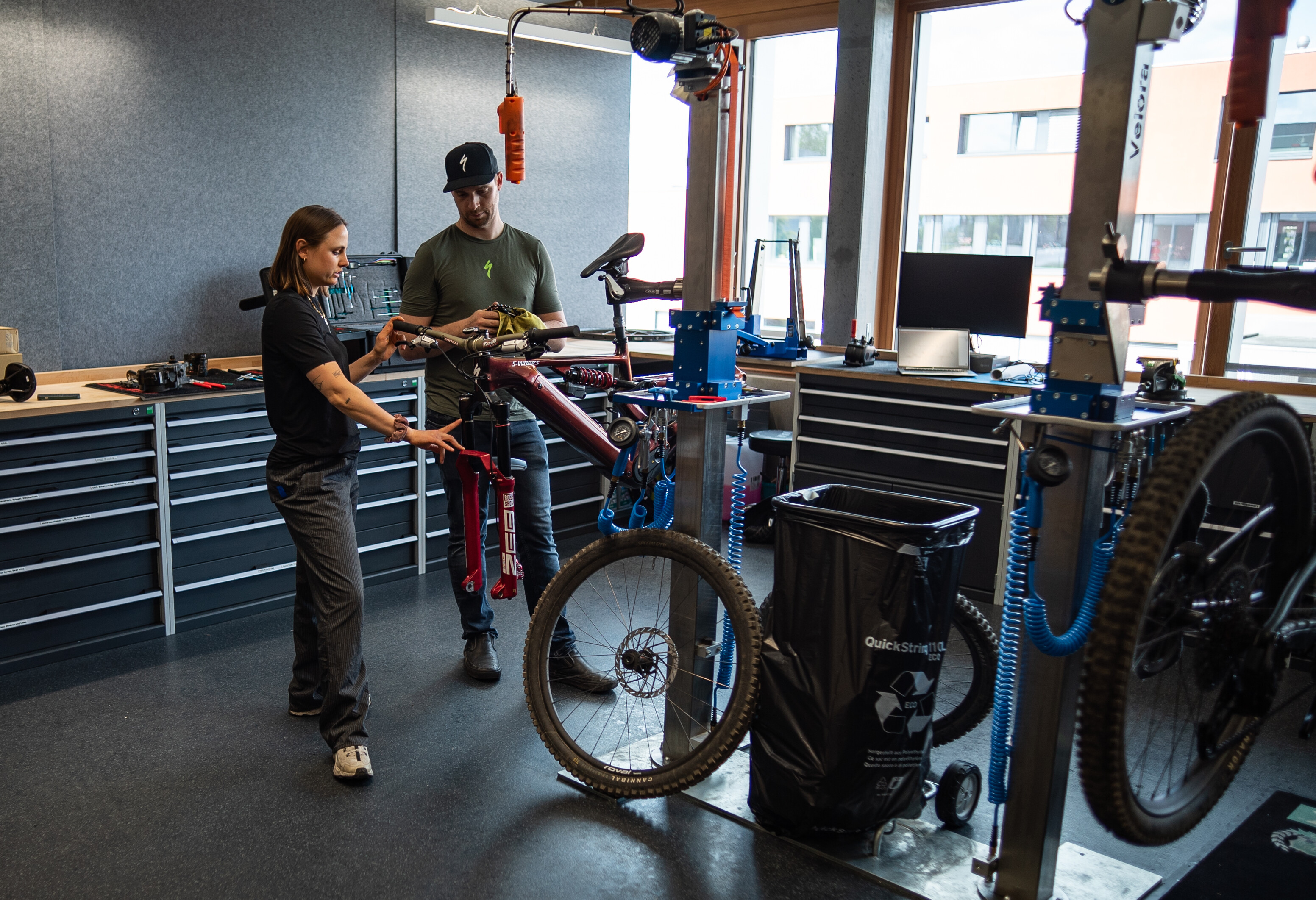 Zwei Personen stehen in einer Werkstatt an einem montierten Fahrrad. Eine Frau zeigt auf ein Bauteil, während ein Mann danebensteht und etwas in den Händen hält.